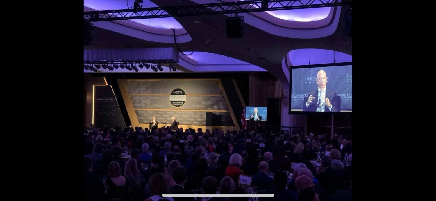 Conference audience facing a speaker and a large screen showing the presenter
